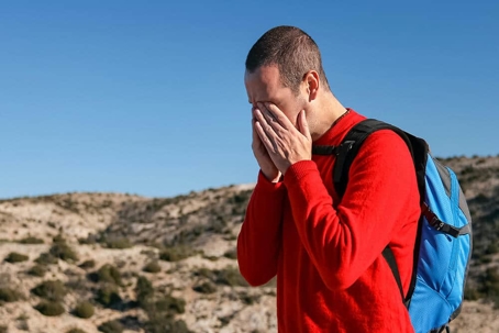 hiker in a red shirt with a blue backpack wipes his eyes
