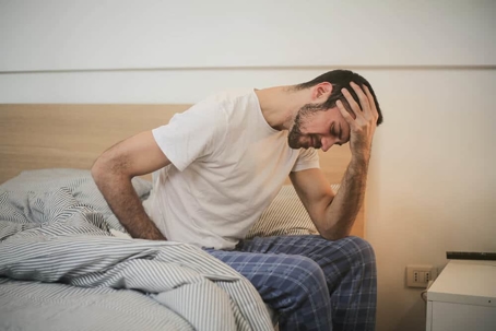 man in pajamas leans in pain on the side of his bed